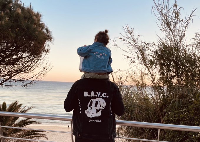 A person stands on a balcony overlooking the ocean at sunset, carrying a child on their shoulders. The adult wears a black jacket displaying "B.A.Y.C." and a "Yacht Club" logo with the iconic bored ape skull design, visually celebrating their membership in the Bored Ape Yacht Club community.