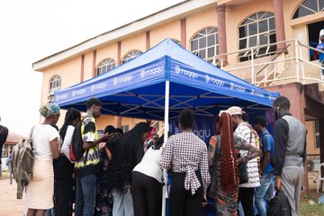 A crowd of people gathered under a blue "ttimes" branded tent outside a beige building, possibly at an outdoor market or event.