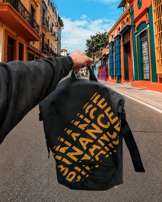 A hand holding a black backpack with yellow text patterns on a colorful street with vibrant orange, blue, and yellow buildings in the background.