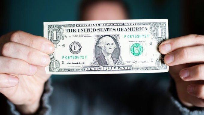 A person holds up a US one-dollar bill featuring George Washington's portrait and the serial number F08759472P, obscuring their face. The image visually represents the "$1.00 bill" referenced in the post, symbolizing the low entry price of buying XRP and the long-term investment potential the author is promoting.