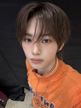 The image features a young man with short hair wearing an orange shirt, standing against a black background. He has his head tilted slightly to one side as he looks directly at the camera. The caption of the image reads "#ALPHADRIVEONE #ALD1 #알파드라이브원 #ANXIN #안신".