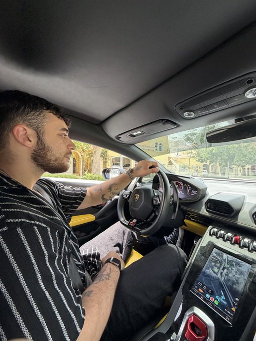 A young man with a beard and tattoos sits behind the wheel of a Lamborghini, identified by the iconic bull logo on the steering wheel, driving through what appears to be a sunny residential area. The luxury supercar's modern interior, featuring a digital dashboard and touchscreen display, visually reinforces the post's "GM" reference, likely highlighting a lifestyle of wealth and automotive extravagance.