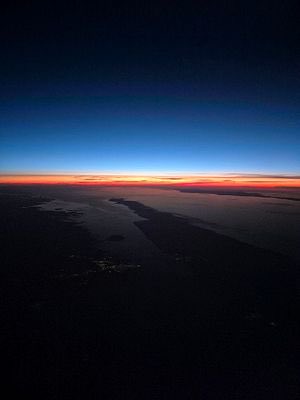 A breathtaking aerial view of a sunrise over the horizon, with the sky transitioning from deep blue at the top to a warm orange glow at the cloud line, overlooking a dark expanse of water and land below.