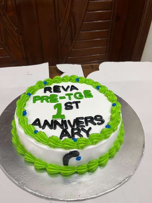 A round white cake with green icing borders sits on a silver board, decorated with the message "REVA PRE-TGE 1ST ANNIVERSARY" in black and green lettering, perfectly capturing the playful celebration of the post marking one year since $REVA's Pre-TGE on 14 August 2025.