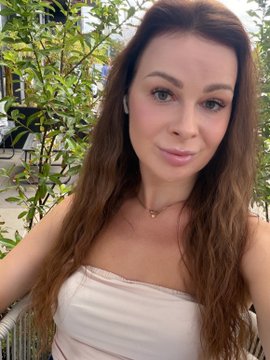 A selfie of a woman with long brown hair, wearing a white top and a delicate necklace, standing outdoors with greenery and a parked car visible behind her.