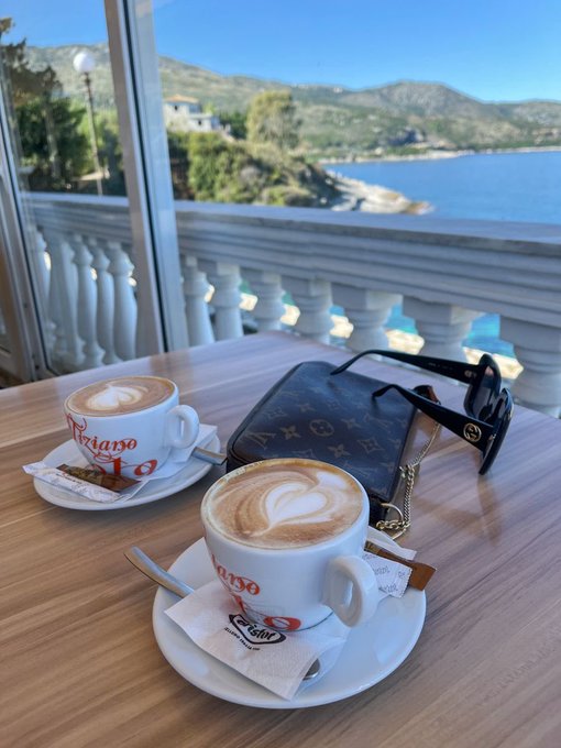 Two cappuccinos in 'Triago' branded cups sit on a wooden table on a balcony overlooking a scenic coastal view of blue water and hills, accompanied by a Louis Vuitton bag.