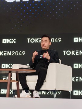 A man in a dark suit sits in a white chair speaking at a TOKEN2049 event, gesturing with his hand during a panel discussion.