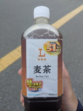 In this photo, there's a closeup image of a hand holding a delicious bottle of barley tea. This closeup image shows Asian characters (Chinese or Japanese) with the translation "Barley Tea" written beneath. Other text includes clues to its purpose, including offering a large 950ml bottle of barley tea for the same price as their smaller 600ml bottles.