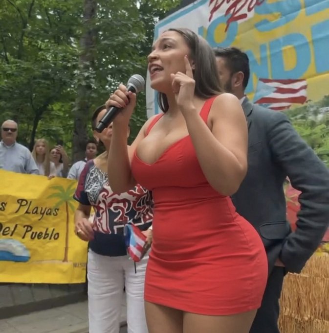A woman in a red dress speaks into a microphone at what appears to be a Puerto Rican parade, gesturing near her head in a pose often associated with communicating "listen to me" or "it's all in the mind." The image appears unrelated to the post about Saylor's USD reserve for $MSTR/BTC.