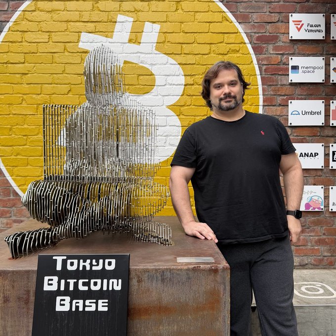 A bearded man in a black t-shirt poses next to a metal Bitcoin sculpture at the "Tokyo Bitcoin Base," with a large Bitcoin logo painted on the yellow brick wall behind him and sponsor logos (including Fulgur Ventures, Mempool.space, Umbrel, and Anapay) displayed on the adjacent wall. The image supports the post's claim that Mark Karpelès, former Mt. Gox CEO (@MagicalTux), visited the Tokyo Bitcoin Base, as he is pictured there alongside the iconic Bitcoin signage.