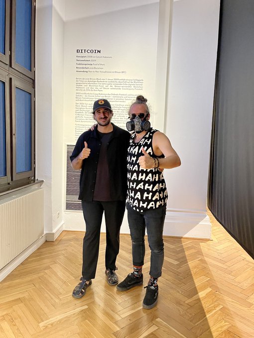 Two men pose giving thumbs up in front of an informational placard titled "BITCOIN" at the Francisco Carolinum museum, with one wearing a "HAHAHAHA" tank top and the other a black cap, likely documenting their meeting at the cryptoart group exhibition mentioned in the post.