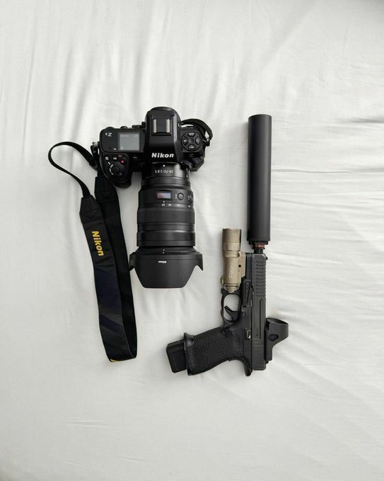 A flat-lay photograph showing a Nikon Z9 camera with a lens and strap next to a black handgun equipped with a suppressor and tactical light on a white sheet.