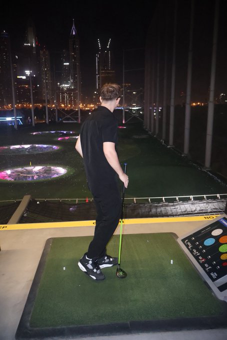 A man in a black t-shirt and sneakers is teeing off at a Topgolf hitting bay at night, with the illuminated Dubai skyline visible in the background. The electronic scoring console on the right shows game options, connecting to the "GN" (Topgolf Night) promotional post.