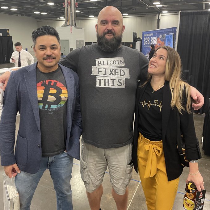 Three people pose together at what appears to be a convention; the man in the center wears a shirt reading 'Bitcoin Fixed This,' while the man on the left sports a Bitcoin logo t-shirt.