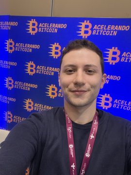 A selfie of a smiling young man wearing a lanyard in front of a blue backdrop covered with 'Acelerando Bitcoin' logos and Bitcoin symbols.
