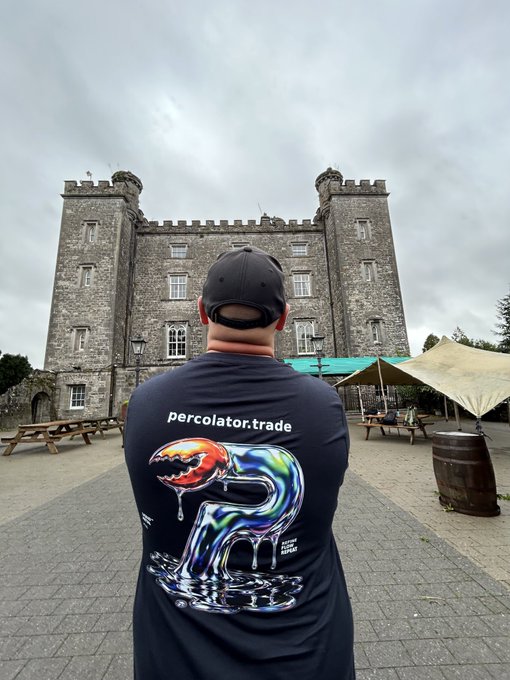 A person wearing a black cap and a black t-shirt featuring 'percolator.trade' and a colorful graphic of a melting figure with a sun stands with their back to the camera, facing a large stone castle or manor building with crenellated towers.
