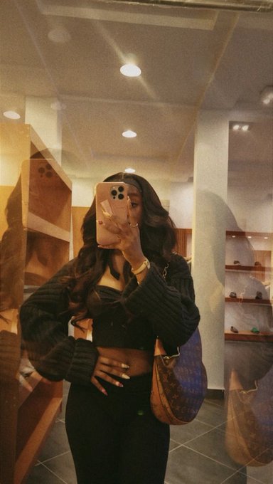A woman takes a mirror selfie with her phone, wearing a black cardigan and carrying a blue quilted handbag.