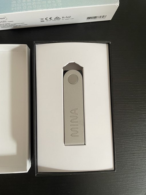 A silver USB drive branded "MINA" sits in a white and black product box on a dark surface, with packaging showing regulatory marks like CE and R-NZ.
