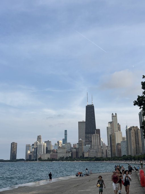 A scenic view of the Chicago skyline from a beach, featuring the John Hancock Center and other skyscrapers against a partly cloudy sky, with people walking along the shore.