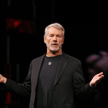 A man with gray hair and a beard, wearing a black blazer over a dark t-shirt, stands on a dark stage with his arms slightly outstretched.