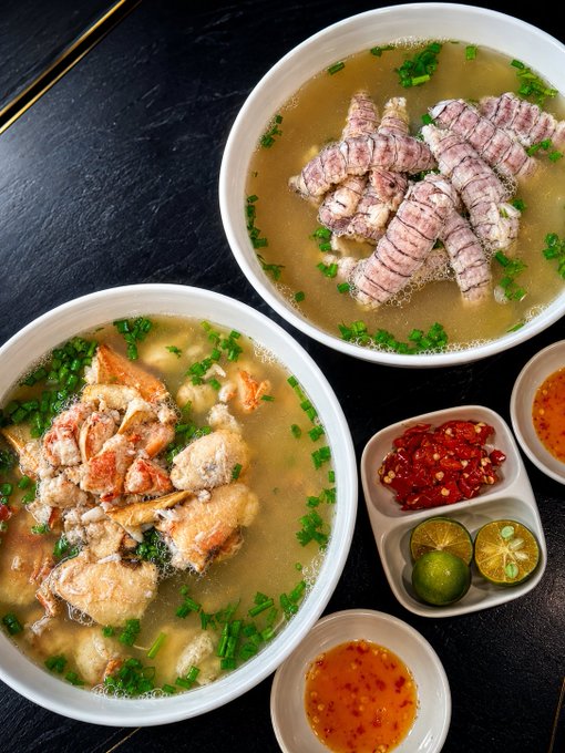 Two bowls of clear broth noodle soup are served, one topped with sliced pork and the other with fried fish, accompanied by small dishes of sliced chilies, calamansi, and chili dipping sauces.