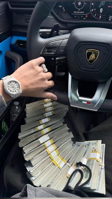 A first-person view from inside a Lamborghini shows a hand resting near the steering wheel, wearing a diamond watch, with a massive stack of banded cash piled on the passenger seat.