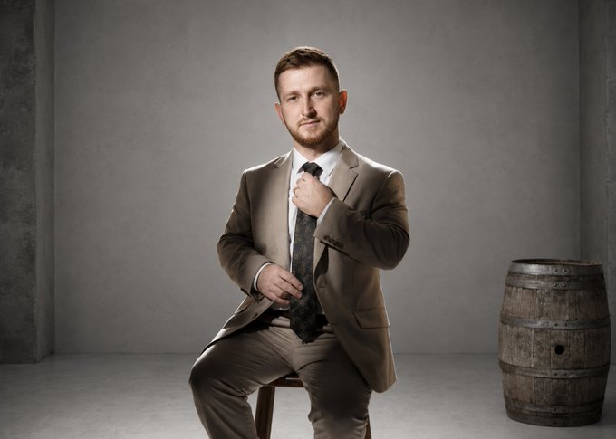 A man in a tan suit adjusts his patterned tie while seated on a wooden stool, with a weathered wooden barrel beside him against a muted gray backdrop—presenting himself as the person running the account.