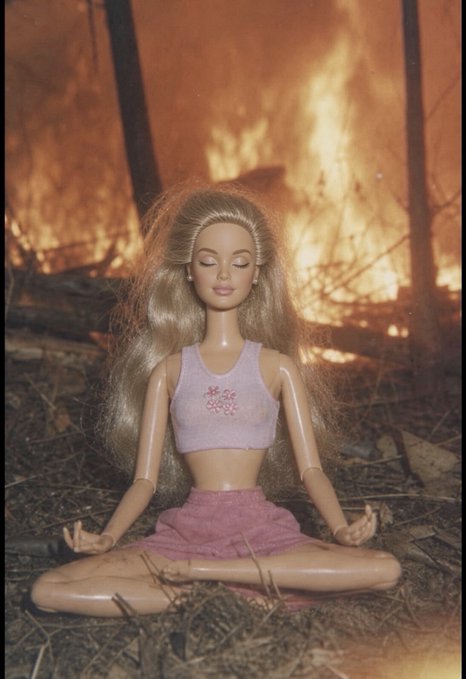 A Barbie doll with long blonde hair wearing a pink crop top and skirt, posed in a meditation position in front of a burning fire.