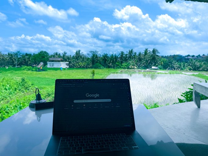 A laptop displaying the Google homepage sits on a glass table overlooking a scenic view of green rice paddies and palm trees under a cloudy blue sky.