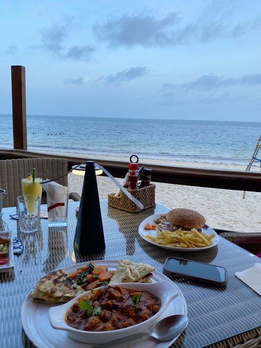 A beachside dinner setting featuring a plate of curry, flatbread, and a burger with fries alongside a lemonade, all overlooking the ocean at dusk.