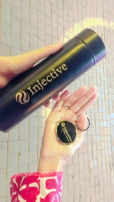 A hand holding a matte black tumbler branded with the Injective logo, alongside a matching Injective keychain, set against a tiled background.
