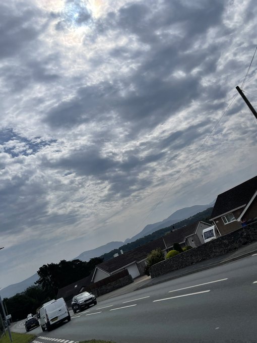 A tilted-angle photograph of a cloudy sky over a residential street with houses, mountains in the background, and vehicles including a white van on the road.