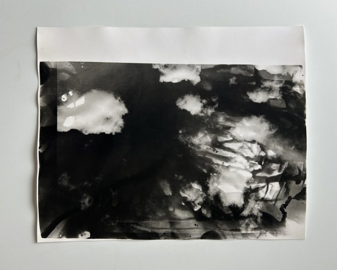 A black and white abstract artwork on paper featuring cloudy, ink-wash style patterns with varying shades of black, white, and gray.