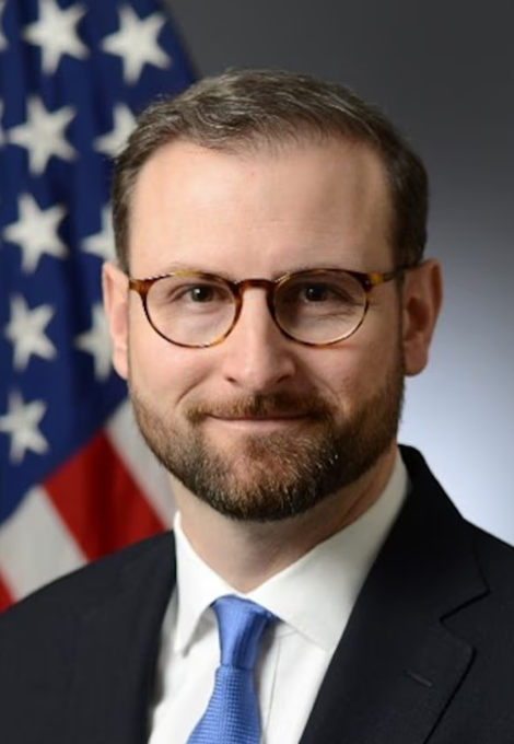 A man with short brown hair, a beard, and tortoiseshell glasses, wearing a dark suit, white shirt, and light blue tie, poses in front of an American flag. The image appears to be an official portrait accompanying a story about White House crypto advisor Patrick Witt's comments on the Clarity Act ahead of a Senate vote.