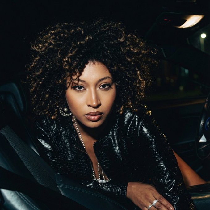 A woman with curly hair and a black leather jacket sits in a car at night, wearing pearl jewelry.