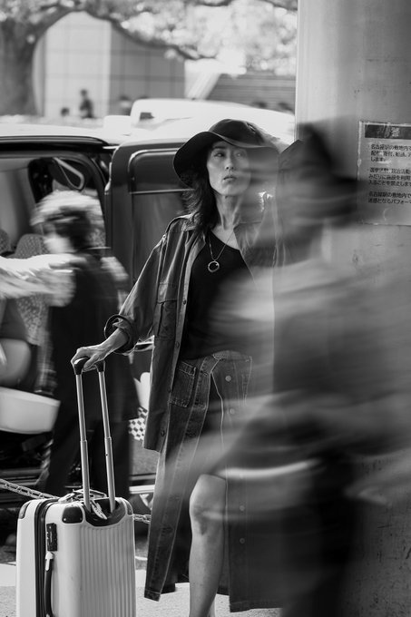 A black-and-white photo shows a stylish woman in a wide-brimmed hat and denim skirt holding a white suitcase, with motion-blurred pedestrians passing by.