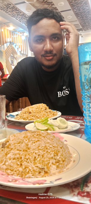A man at a restaurant table in Shaistaganj posing with plates of fried rice and cucumber slices, taken on a realme C75 on August 25, 2025.