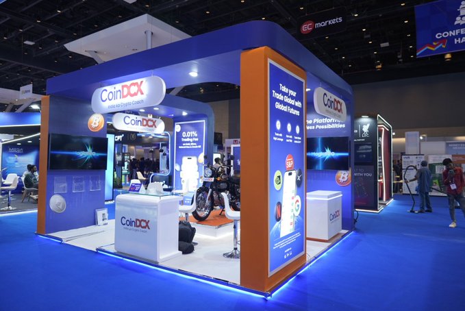 A CoinDCX booth at the Mumbai Money Expo features their branding prominently, displaying promotional text including "Trade your Trade-Bot with Global Futures," "0.01% Trading Fee," and "No Platform Fees, No Possibilities" on illuminated panels. The central display showcases a Royal Enfield Bullet 350 Battalion Black motorcycle, positioned as the giveaway prize for attendees who make a trade at the booth, matching the post's promotion of a contest to win the bike with the winner announcement scheduled for the next day.
