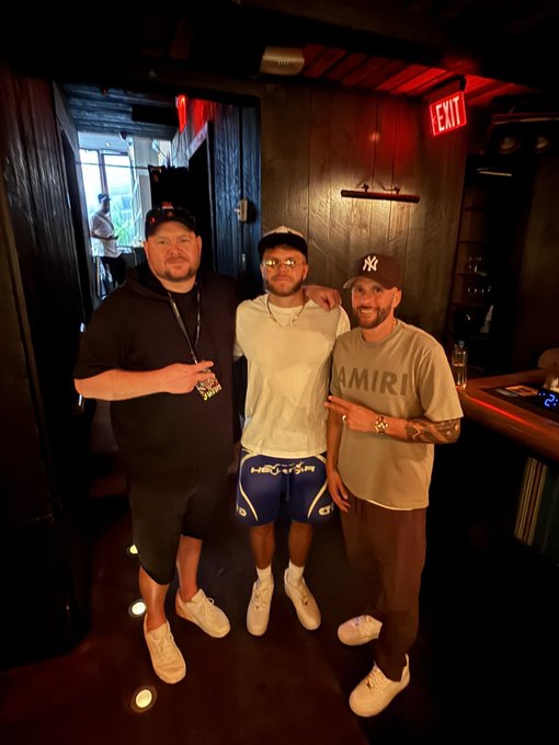 Three men posing together in a dimly lit venue with red lighting; one wears a black hoodie, the middle one wears a white t-shirt and blue boxing shorts, and the third wears a beige 'AMIRI' t-shirt and a New York Yankees cap.