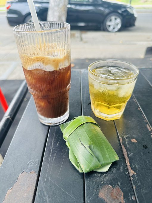Two drinks sit on a weathered black outdoor table—a tall iced coffee and a glass of iced tea—next to a food item wrapped in banana leaves, with parked cars visible in the background.