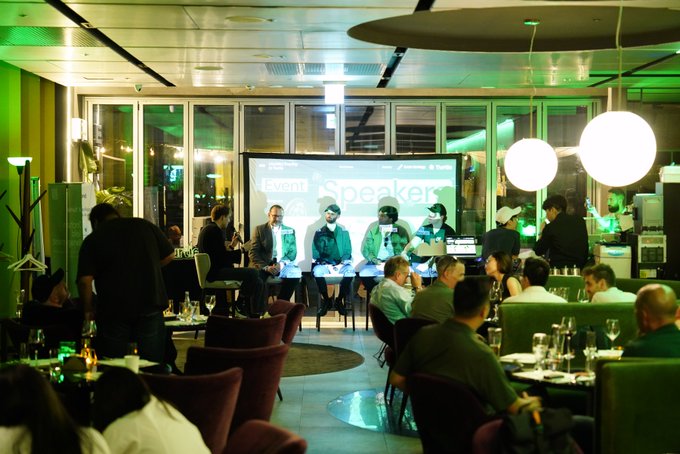 An indoor venue with green lighting hosts a speaker panel discussion with 'Speakers' displayed on a screen, while attendees dine at tables.