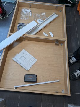 An overhead view of wooden drawers containing various hardware items including white metal bars, plastic packaging, and a small electronic device.