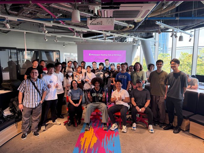 A group photo of approximately 25 people gathered in a modern office or event space, posing for the camera with a projector screen behind them.