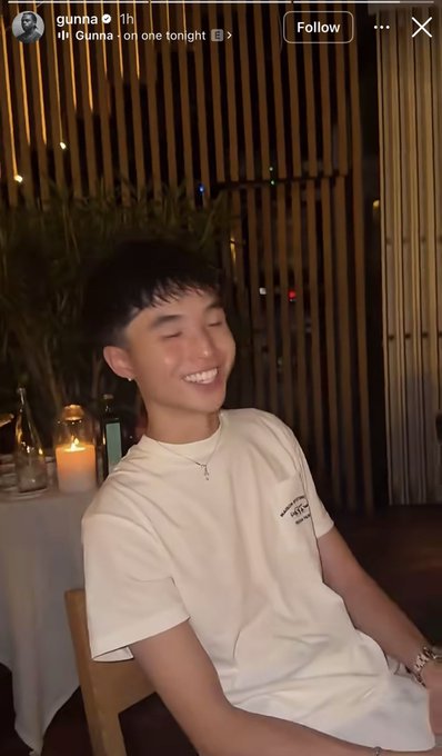 A young man with dark hair and a silver cross necklace is smiling brightly while seated at a dimly lit restaurant with candles and bamboo accents in the background. The Instagram post is from user "gunna" and reads "Gunna on one tonight 🌙". The accompanying caption humorously notes that "Rappers want to be traders, Influencers want to be traders, Kids want to be traders, Memecoins are coming back, Maybe Orangie was right" — suggesting this image of the rapper Gunna ties into the recent trend of celebrities entering the crypto trading space.