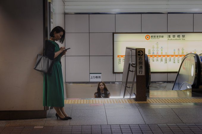 A woman in a green dress and black cardigan stands on a subway platform looking at her phone, while another woman emerges from an escalator at Asakusabashi Station in Tokyo, as indicated by the orange and white line map on the wall.