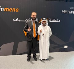 Two men, one in a dark suit with an orange tie and another in traditional white Arab attire, pose together at the Bitcoin MENA conference, with visible signage reading "inmena" and "META P" along with Arabic text in the background. The image reflects the context of the Bitcoin MENA Conference in Abu Dhabi, which has been rescheduled to winter 2027 to ensure a comprehensive experience for the global Bitcoin community.