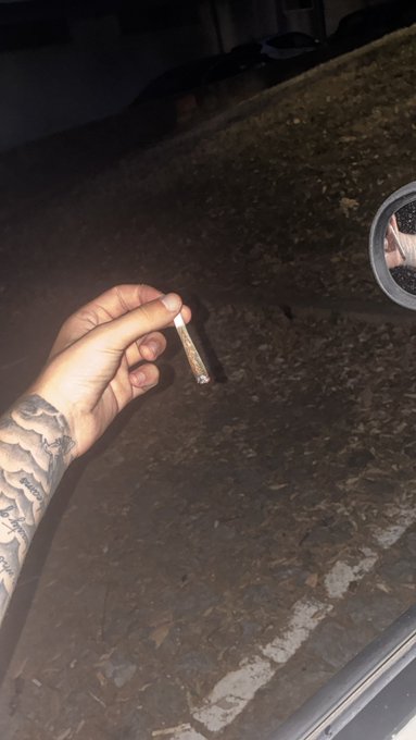 A nighttime photo of a tattooed hand holding a rolled cigarette (joint) outside, with a side mirror reflecting part of the scene and a parked car visible in the background.