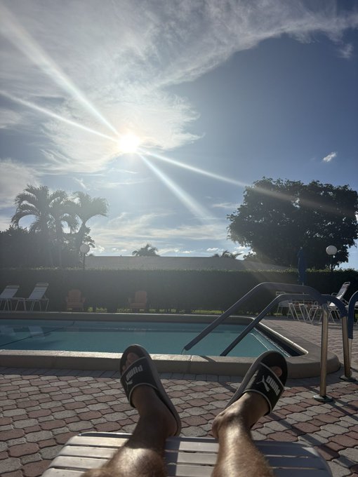 A first-person view of someone wearing Puma slide sandals resting their feet on a lounge chair beside a swimming pool, with the sun shining brightly in a partly cloudy sky above palm trees.