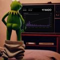 A small green alien figurine stands on a stack of items in front of a television displaying a stock chart for the ticker 'YMAG' with a rising line graph.