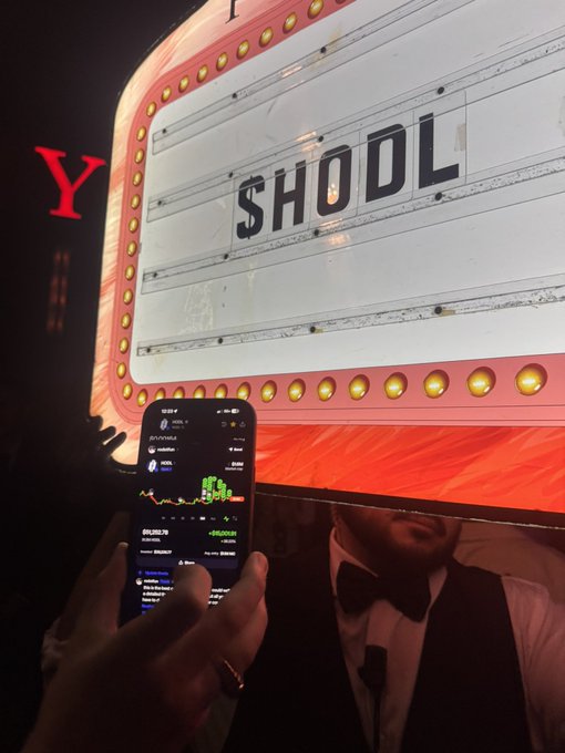 A person in a tuxedo holding a smartphone displaying a stock trading chart with green numbers, standing in front of a large illuminated 'SHOOL' marquee sign.
